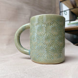 taza de cerámica hecha a mano con esmalte verde brillante y chipas celestes y relieve de margaritas.