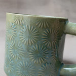 taza de cerámica hecha a mano con esmalte verde brillante y chipas turquesas y relieve de margaritas.